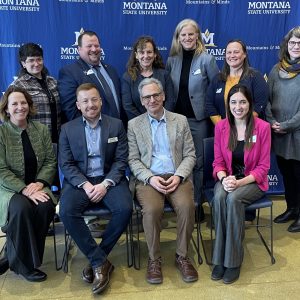 Two rows of people (one sitting and one standing) smiling in front of a blue Montana State University backdrop.