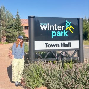 Ava Fricke, wearing a Winter Park cap and a striped blue shirt, stands beside the Town of Winter Park Town Hall sign on a sunny day.
